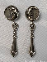 Pair of sterling silver vintage moon face earrings with dark patina displayed flat on fabric. Shows front view of the earring pair illustrating the round moon face and the long drop pendant.