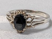 Close-up photo of the sterling silver ring with an oval dark blue sapphire set in a prong setting with decorative silver bead accents, showing the front and side view of the band.