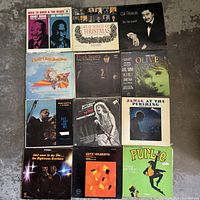 Photo showing 12 vintage vinyl record albums arranged on a floor. Album covers are visible with titles and artist names, variety of genres apparent.