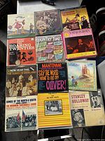 A wide view of 11 vintage vinyl records laid out on a flat surface. Covers include The Sound of Music, Clancy Brothers Greatest Hits, Music from the Roaring Twenties, Country Times, A Star Is Born, and more.