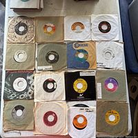 Overview of 16 vintage 7-inch vinyl records on a flat surface showing worn paper sleeves, various labels including ABC Paramount and MGM.