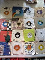 Overview photo showing 16 vintage 7-inch vinyl records with various colored labels and paper sleeves, some with original covers.