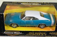 Front and side view of boxed 1969 Dodge Charger R/T 1:18 scale die cast model. Blue body with white roof and stripes, visible chrome wheels, framed in clear plastic box with yellow background.
