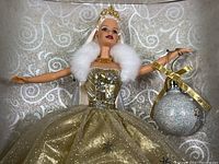 Close-up of Barbie doll's upper body showing white faux fur stole, gold crown and necklace, holding a glitter ornament marked 2000.