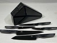 Full set of five black Dalstrong Shadow Black Series knives laid out next to black triangular block on white surface.