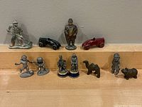 Miniature antique metal toys including human figurines, toy race cars, and animals displayed on a wooden ledge against a gray wall.
