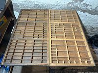 Two wooden antique printing trays placed on the floor against a wall. Each tray has multiple compartments in various sizes used for holding type keys in printing.