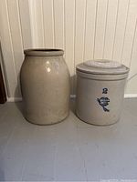 View of two stoneware crocks side by side; a larger plain crock and a smaller lidded crock with blue decoration and markings.
