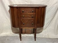 Front view of closed antique walnut sewing cabinet showing three drawers with brass knobs and two closed side drop-leaf doors.