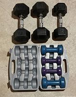 Photo showing three pairs of hex rubber dumbbells with chrome handles (two 25 lb pairs, one 15 lb pair) and a case holding six vinyl coated dumbbells in blue and purple ranging from 3 to 8 lb.