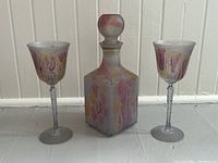 Front view of the decanter between two matching goblets, showing the soft lilac and pastel color patterns, frosted glass bases, and spherical stopper on the square decanter.