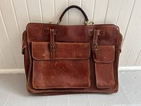 Front view of brown leather briefcase showing two front buckle flap pockets, leather texture with wear and patina, top handle.