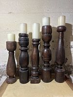 Set of five wooden candlesticks of varied shapes and sizes, each holding a white candle, displayed against a light wall.
