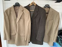 Three men's sports jackets hanging on a rod, shown front side. Beige jacket on left, dark brown houndstooth jacket in center, beige Ralph Lauren jacket on right.