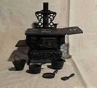 Front view of miniature antique cast iron Crescent stove with four burner plates on top, fold-out side shelf, and two small handles projecting. Includes seven cast iron miniature cooking accessories set in front: bucket, pots, frying pan, spatula.