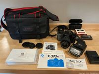 Full kit overview showing camera with attached lens, flash, Blacks carrying case, manuals, lens caps, battery charger, and film canisters.