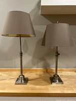 Pair of silver column lamps on wooden surface with gray fabric shades. The lamp on the left is taller than the lamp on the right.