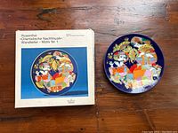 Photo shows the Rosenthal porcelain wall plate alongside its original box with matching artwork and labeling. The plate's colorful and detailed design is clearly visible.