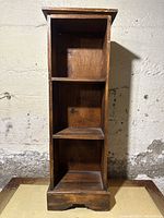 Front view of a small wooden shelf with three compartments and carved base against a concrete wall.