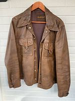 Full front view of vintage brown leather jacket hanging on wooden hanger showing snap close front and chest pockets.