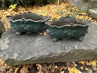 Pair of small antique cast iron planters shown outdoors on stone surface with fall leaves in background.
