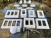 Various white plastic wall switch plates including single, double, triple, and quadruple configurations laid out on a glass table outdoors, along with a construction bulb.