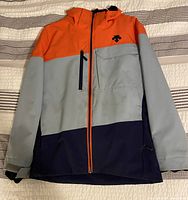 Front view of Descente youth ski jacket showing color block design in orange, grey, and navy blue with zippered front and adjustable cuffs.