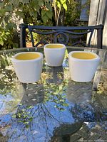 Photo showing three white ceramic egg cups with yellow interior placed on a glass table against an outdoor background.
