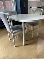 Angle view of the oval white kitchen table with two of the matching chairs positioned around it, showing table legs and chair backs with light upholstery and minor wear visible.