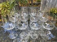 Set of twelve clear Reidel Cabernet wine glasses displayed outdoors on a glass table with garden background.