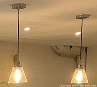 Close-up photo showing one installed pendant light lit with visible light bulb, highlighting the square ribbed frosted glass shade with black fabric cord.