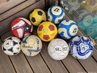 Nine soccer balls on a wooden floor including inflated and foam types.