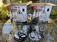 Two unpacked Globe Electric Parker Edison-style pendant lights with clear glass shades, chrome fixtures, and cords coiled, shown with their retail boxes in the background outdoors on a glass table.