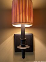 Close-up of one wall sconce mounted on wall, showing pleated ivory shade lit, matte black square base and cylindrical arm.