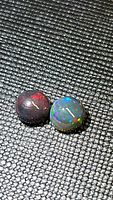 Close-up photo of two round cabochon black opal gemstones on textured dark background, showing their characteristic rolling rainbow flashes in red, blue, and green hues.