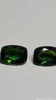 Two rectangular cushion-cut green gemstones displayed side by side on a white background, showing deep electric green color and clarity.