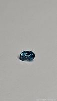 Oval cut natural zircon gemstone with deep lagoon blue color shown on neutral background under lighting to highlight color and clarity.