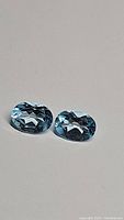 Pair of natural oval cut sky blue topaz gemstones on a plain surface, showing their size, shape and color clearly.
