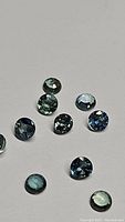 Clear close-up image of ten round cut sapphires varying in teal blue and green tones placed on a gray surface.