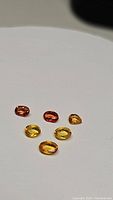 Six faceted Ceylon sapphires in mixed deep orange and yellow hues laid out on a neutral background, showing size and color variation.