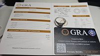 Photo showing GRA grading report indicating 2ct carat weight, D color, VVS1 clarity, excellent polish and symmetry. Ring next to certificate.
