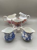 Five assorted creamers arranged showing different patterns and conditions, including blue and white Royal Crown Derby and Imari style pieces