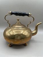 Front and side view of brass teapot showing its round shape, black wooden handle, spout, and three round feet.