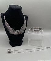 Three sterling silver pieces displayed on bust and block: multi-chain necklace, ball locket with chain, engraved bangle bracelet.
