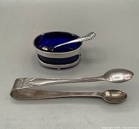 Photo showing the cobalt blue glass liner inside the sterling silver salt cellar, gold washed salt spoon placed inside the liner, and sterling silver tongs in front.