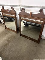 Full view of the two wooden framed vanity mirrors standing upright, showing the carved decorative details, fretwork panel, and beveled glass mirrors.