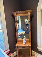 Tall rustic wooden table with attached large rectangular mirror and four double hooks on the mirror frame. Drawer below the tabletop with a handle. Photo shows the furniture piece in a corner next to windows.