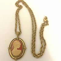 Front view of an oval pendant necklace with a faux cameo depicting a lady's profile in cream on an orange background, set in textured gold tone metal with a matching gold tone rope chain and lobster clasp.