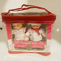 Two Campbell Kids dolls in original packaging plastic bag with orange zipper and handle, showing dolls dressed as chefs with a packaging card behind them.