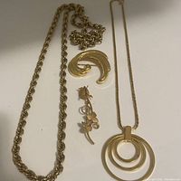 Photo showing gold tone twisted rope necklace, curved brooch, rose pin, and necklace with triple circle pendant on white background.